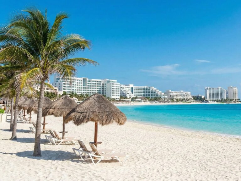 Sunny Cancun Hotel Zone beach with white sand, turquoise Caribbean water, palm trees, and luxury beachfront resorts in the background. Cancun Hotel Zone. luxury resorts